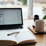 A laptop is on and sitting on top of a light colored desk next to a white coffee mug and a notebook with a pen sitting on top of it.