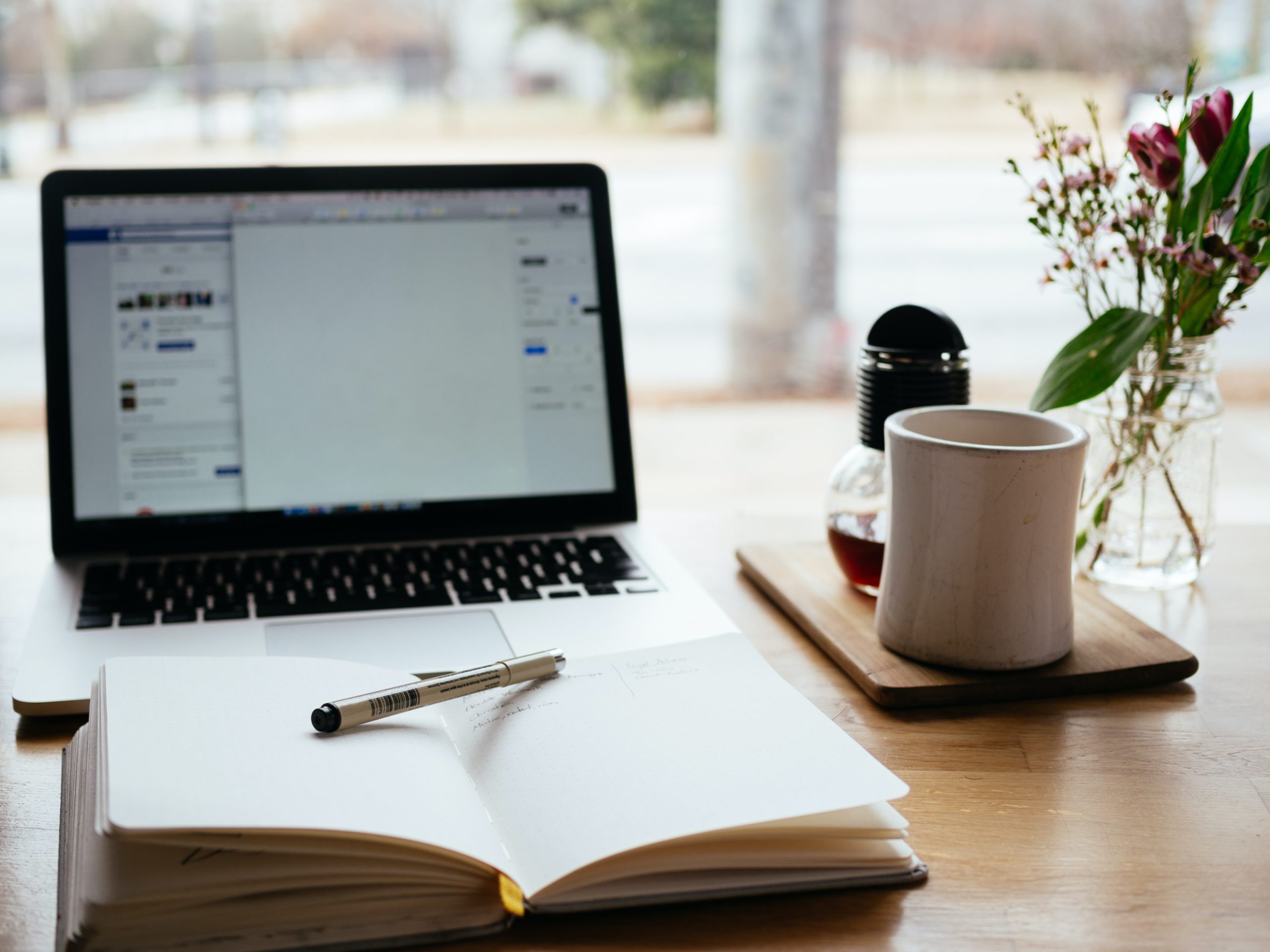 A laptop is on and sitting on top of a light colored desk next to a white coffee mug and a notebook with a pen sitting on top of it.