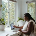 A dark skinned person with long braids interacts with their laptop on the left. Their office is bright with white walls, desk, supplies, and plant pot. Past the windows are relaxing greenery and trees.